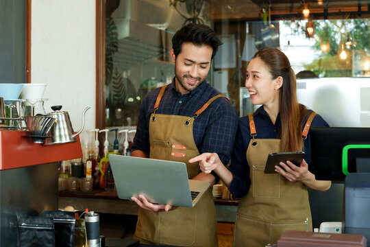 Asian couple runs cafe bakery together, man holding laptop computer, woman holding laptop, different people working in their own coffee shop, woman secretly turned look at man work being done.