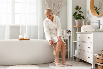 Mature woman massaging her leg with brush in bathroom - obrazy, fototapety, plakaty