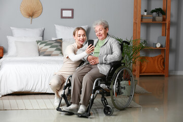 Fototapeta na wymiar Young caregiver with senior woman in wheelchair using mobile phone at home