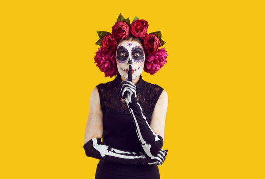 Woman asks to keep her secret on Day of Dead or Halloween. Lady in Catrina costume with sugar skull face and wreath of beautiful roses doing shhh tss sign standing isolated on color studio background