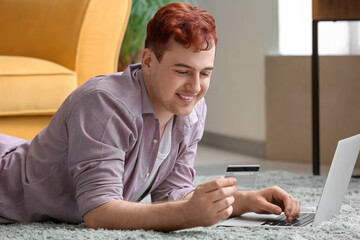 Handsome young man with credit card and modern laptop lying on floor while shopping online