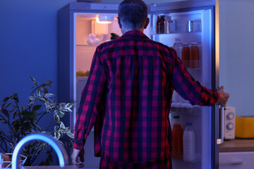 Hungry mature man near open fridge in kitchen at night, back view
