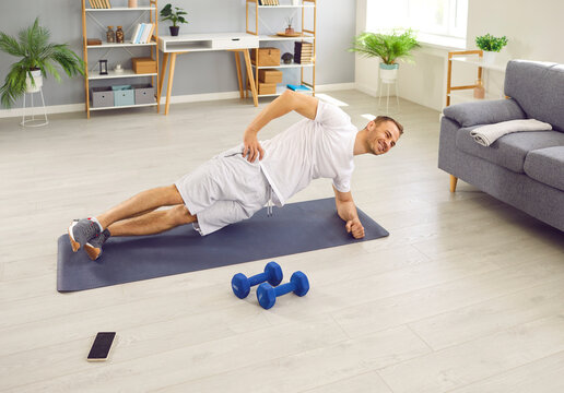 Happy, fit, sporty, young man having a fitness workout at home and doing a side plank exercise on a sports mat on the floor in the living room. Body training, healthy lifestyle concept