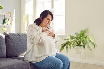 Portrait of a young plump overweight fat woman sitting on the couch in the living room at home putting a hand on her chest suffering from heart problems. Obesity problems and healthcare concept.