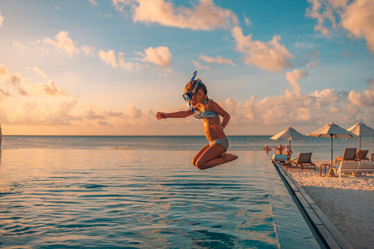 Amazing Sunset Sea Sky With Joyful Girl In Snorkel Gear Jumping Into Pool. Tropical Resort Beach Umbrellas, Infinity Poolside Excited Funny Little Girl Jumping To Swimming Pool. Happy Summer Vacation