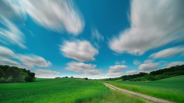 time lapse of summer landscape of a green field with a country road and white clouds floating across the blue sky leaving a trail