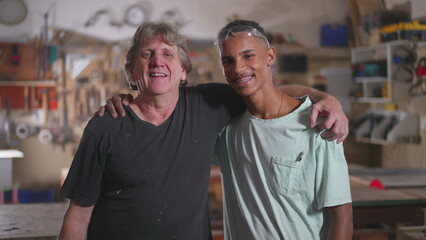 Smiling Senior Carpenter and Apprentice Posing in Workshop. Senior boss putting arm around junior diverse employee standing in carpentry place with tools in background