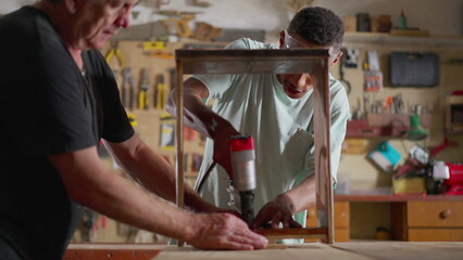 Young carpenter working at workshop with the help of his teacher. Senior man guiding pupil with furniture building, woodworking job occupation