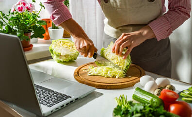 Woman prepares a vegetable salad in the kitchen using laptop computer looking at recipe.