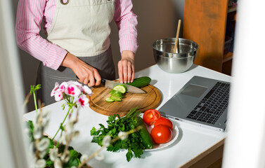 Woman prepares a vegetable salad in the kitchen using laptop computer looking at recipe.