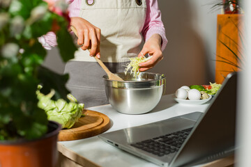 Woman prepares a vegetable salad in the kitchen using laptop computer looking at recipe.