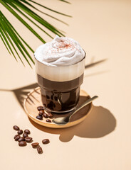 Iced coffee in a glass with foam on a yellow background with coffee beans, palm leaf and  shadows. Summer refreshment concept.