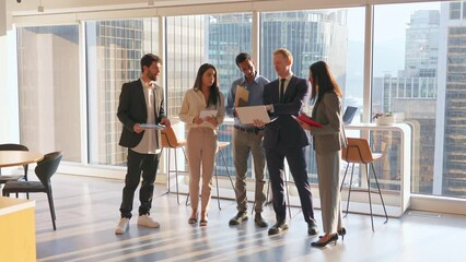 Young happy diverse professional team business people standing in modern office and talking. International workers group having teamwork discussion planning strategy with devices at work meeting.
