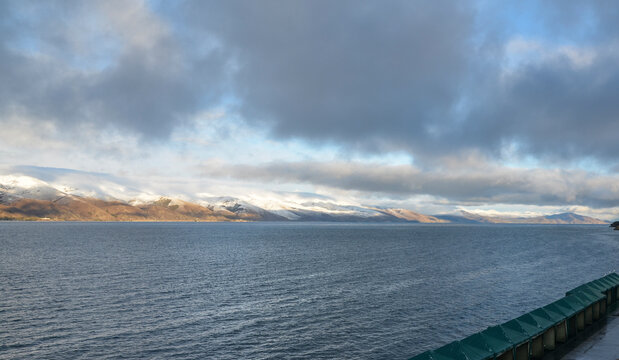 Calm water of Lake Sevan at the foot of the snow-capped mountain peaks under blue sky with low clouds. Amazing natural landscape in Armenia