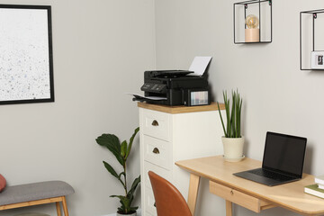 Modern printer with paper on chest of drawers near wooden table indoors
