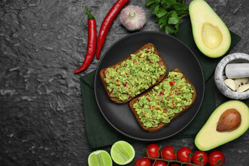 Slices of bread with tasty guacamole and ingredients on black textured table, flat lay. Space for...