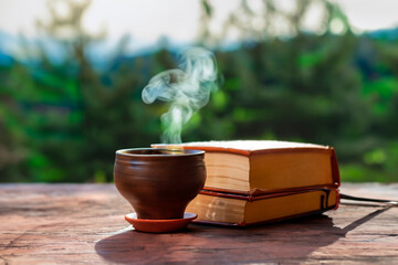 A ceramic cup with hot liquid, next to some books in the background of a rural landscape.