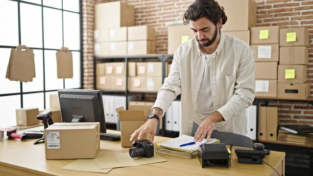 Young hispanic man ecommerce business worker packing professional camera on cardboard box at office