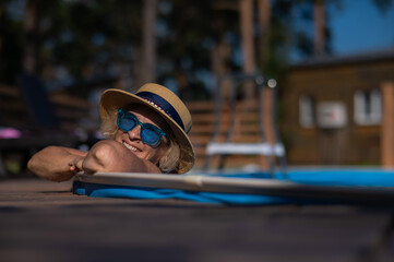 Portrait of an elderly woman in a hat and blue sunglasses swims in the pool. Vacation in retirement. 
