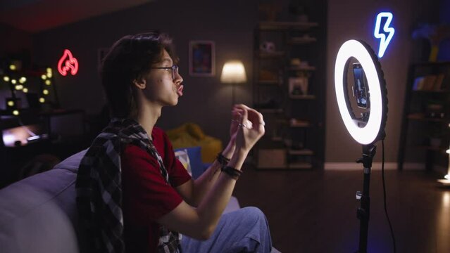 Asian guy with glasses shoots conversational emotional stream while sitting on sofa in front of ring led lamp. Blogger recording video for fun. Concept of contemporary virtual life self-expression 
