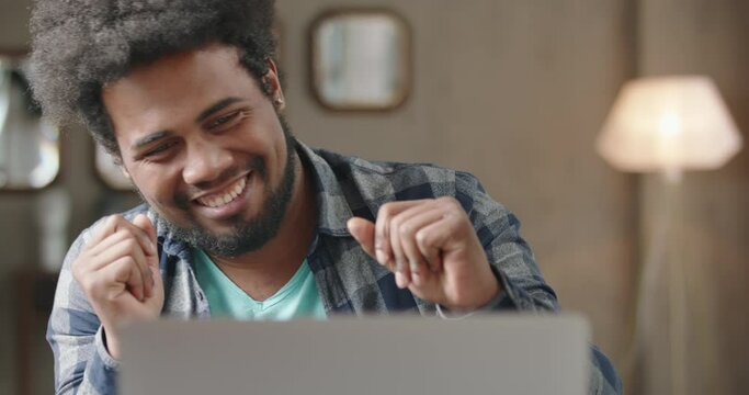 Young black man working on laptop. Man expressively rejoices, laughs, dances merrily in good happy mood. Luck, victory, winning in game. Concept of betting, gambling. Black man wins lottery.