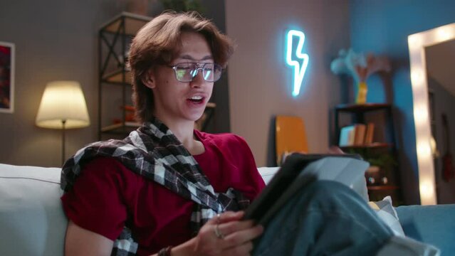 Asian guy in glasses with tablet sits on sofa, online communication. Vibrant expressive emotion. Cinematic. Young man communicates with someone via video link, covers mouth with his hand in surprise
