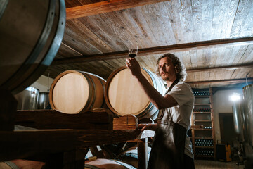 Winemaker tasting wine in a cellar with wooden barrels - obrazy, fototapety, plakaty