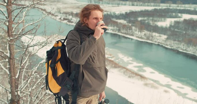 positive young man enjoying drinking hot tea, while standing on the top of mountain Slow motion, traveler has rest during hicking, freedom, happiness, hicker gets pleasure from adventure, river in