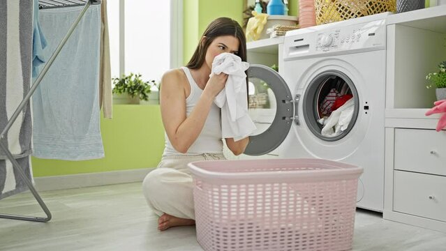 Young beautiful hispanic woman washing clothes smelling dirty clothes at laundry room