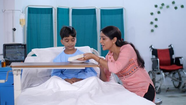 A caring Indian mother is feeding her sick son while in the hospital - care and assistance  healthy food. A small boy is eating his breakfast in the morning while sitting on the hospital bed - a nu...