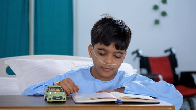 A small kid / an adorable Indian child is reading a book while alone in the hospital - medical treatment  brave boy  hospital care. A cute boy with an oxygen tube is admitted to the hospital - medi...