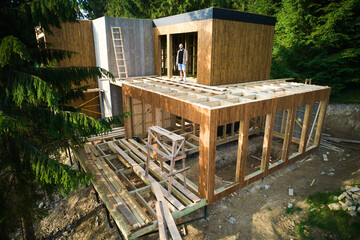 Architect is on construction site of wooden frame two-story house located near the forest. The concept behind it is modern and environmentally friendly construction.