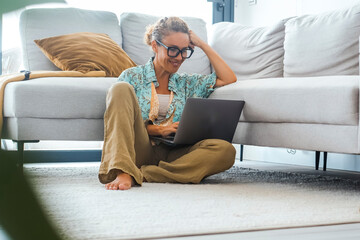 Carefree modern adult woman using laptop at home sitting on the floor against a grey sofa. Female...