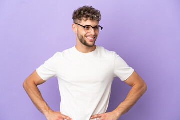 Delivery caucasian man isolated on purple background posing with arms at hip and smiling