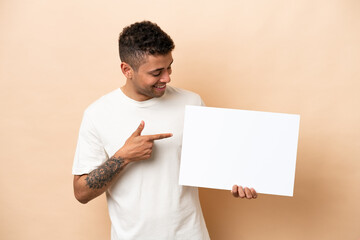 Young Brazilian man isolated on beige background holding an empty placard with happy expression and pointing it