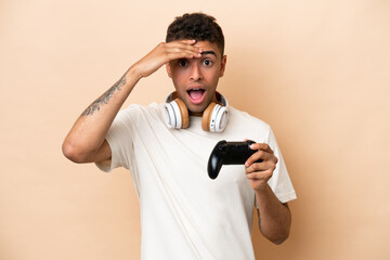 Young Brazilian man playing with a video game controller isolated on beige background with surprise expression