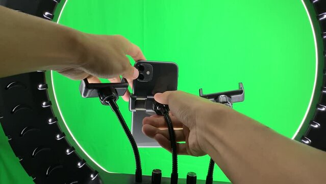 Hand adjusting phone on holder grip on live streaming tripod stand ring light with green screen chroma key background inside photography studio