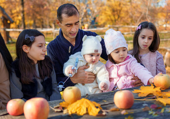 family has picnic in autumn city park, father and daughters, children and parent sitting together at the table, with apples and yellow maple leaves, happy people enjoying beautiful nature