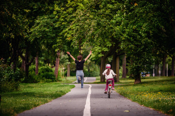 Fototapeta na wymiar Young mother in jeans and black t-shirt running and playing with her daughter in summer parks while girl riding a bike