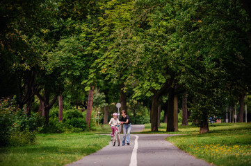 Young mother in jeans and black t-shirt teaching and playing with her daughter in summer parks while girl riding a bike