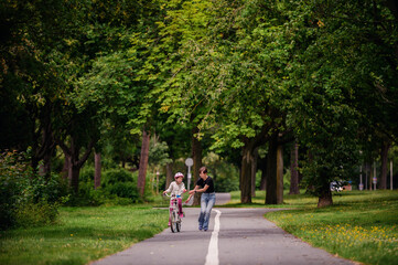 Young mother in jeans and black t-shirt teaching and playing with her daughter in summer parks while girl riding a bike