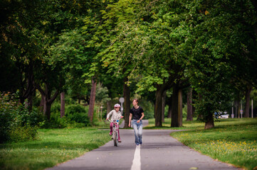 Fototapeta na wymiar Young mother in jeans and black t-shirt teaching and playing with her daughter in summer parks while girl riding a bike