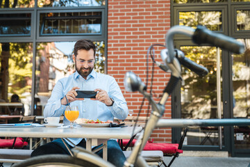 Happy manager sitting in the restaurant outdoors and having a breakfast. Smiling man taking a photo of his food during the sunny day.
