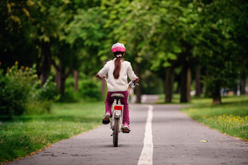 Little schooler girl riding bike in parks. Summer time, wearing helmet. From the back.