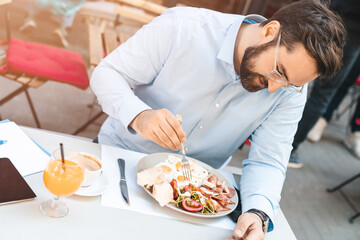 Businessman having a breakfast in fancy restaurant during the sunny day. He is sitting outdoors and looking at his plate with ham and eggs.