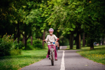 Obraz premium Little schooler girl riding bike in parks. Summer time, wearing helmet. From the back.