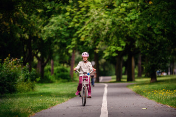 Little schooler girl riding bike in parks. Summer time, wearing helmet. From the back.