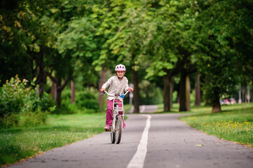 Little schooler girl riding bike in parks. Summer time, wearing helmet. From the back.