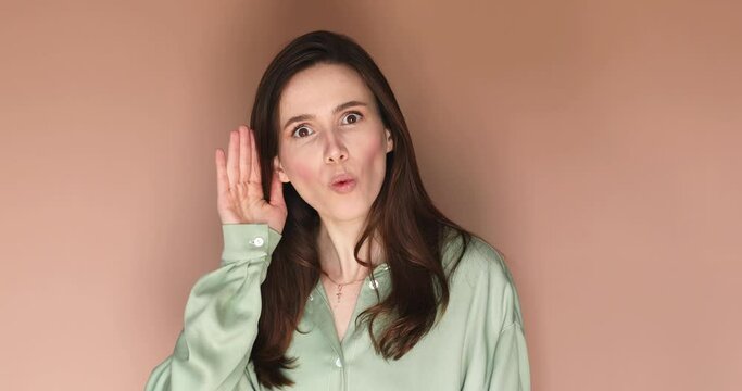 Cute curious woman keeps hand near ear tries to overhear something very interesting listens somebody secret looks camera likes collecting rumors poses against beige background. Eavesdropping concept.