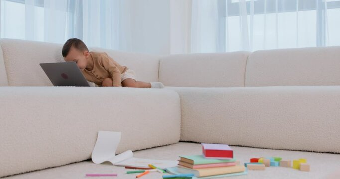 cute curious little boy sitting open notebook, studying online morning, typing, using computer sitting on living room sofa house Funny child send e-mail while mommy is cooking in kitchen Slow motion